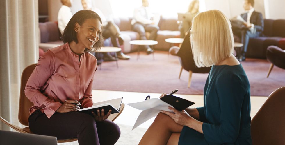 Two professional women building social capital through a friendly business conversation in a modern office lounge.