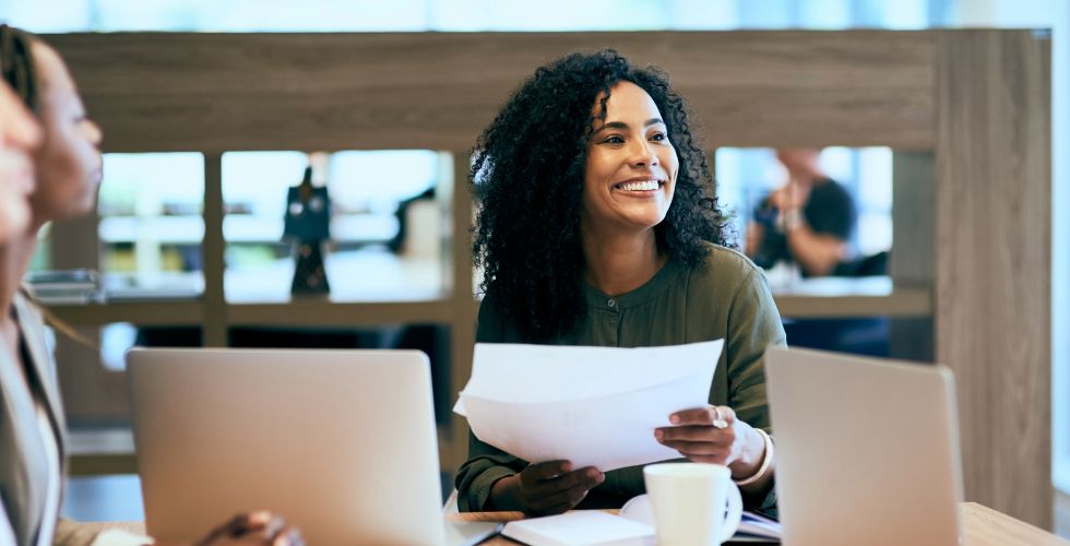 A professional woman smiling during a team meeting with laptops and documents, illustrating the ease of working with a modern data enrichment service.