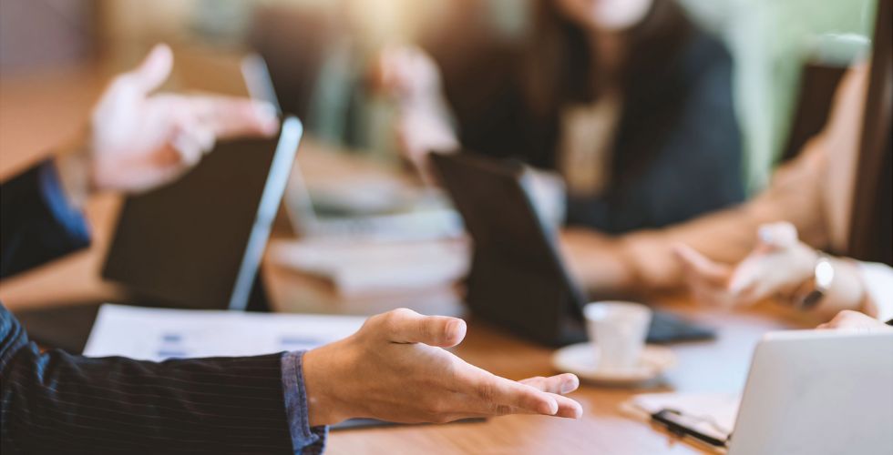 Business professionals seated around a conference table in a collaborative meeting, with laptops, notebooks, and coffee visible, as one person gestures while discussing strategy. The image represents marketing and business development teams using shared relationship data and CRM insights to coordinate outreach, plan growth initiatives, and connect relationships to revenue outcomes in a professional services firm.