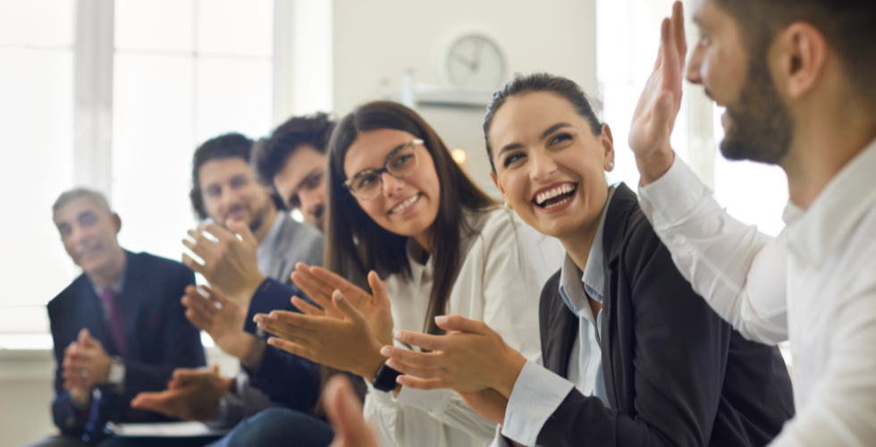A diverse group of business professionals seated in a conference room applaud and exchange smiles during a meeting, representing how search intelligence solutions help firms quickly surface shared relationship context, past interactions, and client insights before important conversations.