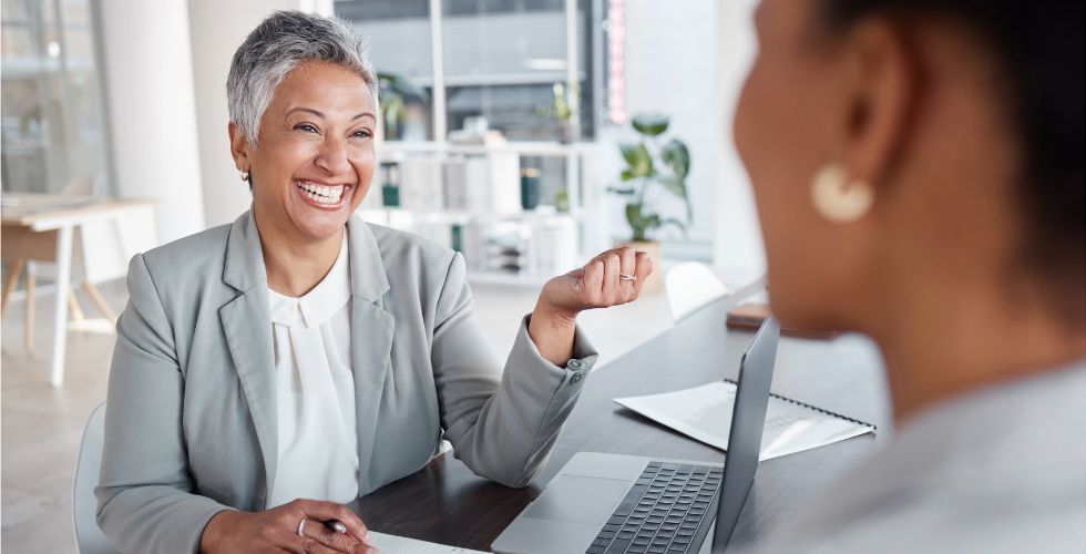 Professional middle-aged woman with short gray hair, smiling and gesturing while engaged in a business conversation with a colleague across the table in a bright, modern office setting. Both are dressed in business attire and seated at a desk with an open laptop, notebook, and documents. The scene suggests a collaborative discussion, reflecting productive CRM enhancement strategies like relationship mapping or improving client insights within existing systems.