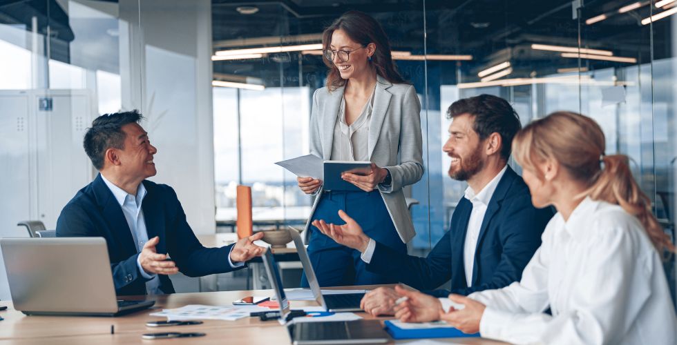 Four business professionals in a modern office meeting reviewing documents and discussing client relationships and contact data, representing enterprise contact management and collaboration across teams.