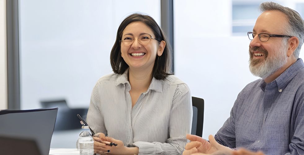 Two business professionals collaborating in a modern office meeting, representing enterprise data management in practice. A woman with glasses and shoulder-length dark hair sits at a conference table with a laptop, pen, and water bottle, smiling as she listens. Beside her, a bearded man wearing glasses gestures while speaking, both appearing engaged in a strategic discussion. The scene reflects teamwork, communication, and relationship-driven decision-making central to enterprise data management and business operations.