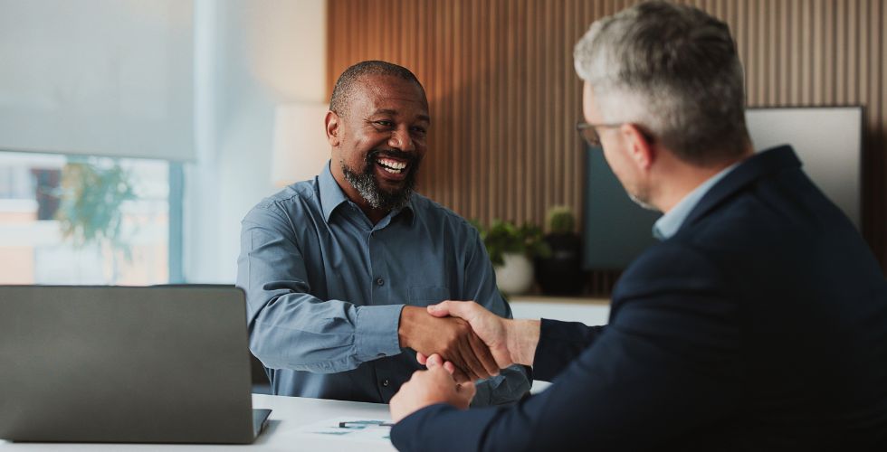 Two business professionals shaking hands across a desk during a meeting, representing managing client relationships through trust, collaboration, and data-driven decision-making in a modern office setting.