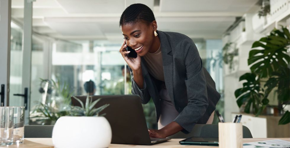 A law firm business development professional in a gray blazer multitasking on a phone call while reviewing law firm business intelligence software on a laptop in a modern, plant-filled office environment, representing the use of legal business intelligence and relational BI tools to manage client relationships and identify growth opportunities in real time.