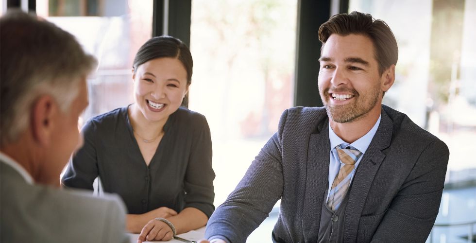 A bright, naturally lit office meeting scene showing three business professionals engaged in a positive face-to-face conversation. An older white man with grey hair is seen from behind in the foreground. Facing him are a smiling East Asian woman in a dark grey blazer and a smiling white man in his thirties wearing a grey suit jacket and a tan striped tie. The setting and body language suggest a senior client meeting or business development conversation — the type of relationship activity that, as the article argues, is rarely captured in CRM records but is exactly what agentic AI systems need to act reliably.