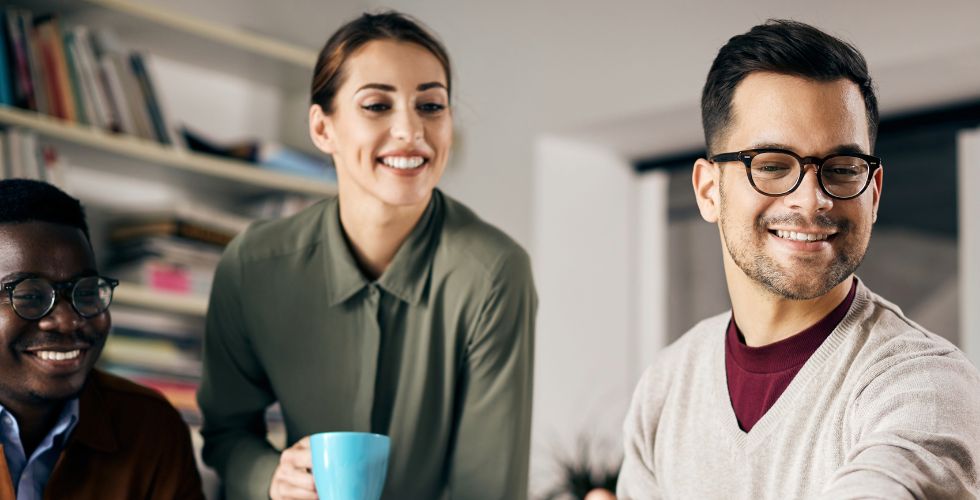 Three professional colleagues in a casual office setting, engaged in a relaxed but focused conversation. In the foreground, a man with short dark hair, glasses, and a light beige v-neck sweater over a burgundy shirt smiles while looking downward, suggesting he is reviewing shared content. To his left, a man with short natural hair and round glasses wears a light blue shirt and is also smiling. Standing behind them, a woman with dark hair pulled back, wearing an olive green blouse, holds a light blue mug and smiles toward the group. A bookshelf filled with books and binders is visible in the background, placing the scene in a professional or office library environment. The mood is collaborative and relaxed, consistent with a pre-meeting or internal briefing context in a professional services firm.